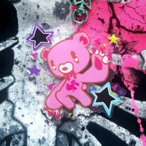 Puede incluir: Llavero de oso rosa con cierre en forma de estrella. El oso tiene un diseño desgastado con salpicaduras de sangre y marcas de garras. Se incluyen dijes adicionales en forma de estrella en negro, morado y amarillo. El fondo presenta un diseño salpicado en rosa, negro y blanco.