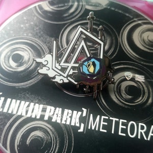 Op de afbeelding: Metalen pin met het Linkin Park-logo, een gestileerd ontwerp van een wolk en een geometrische vorm met een kruik. De pin staat op een zwarte achtergrond met de bandnaam en de albumtitel, "Meteora."