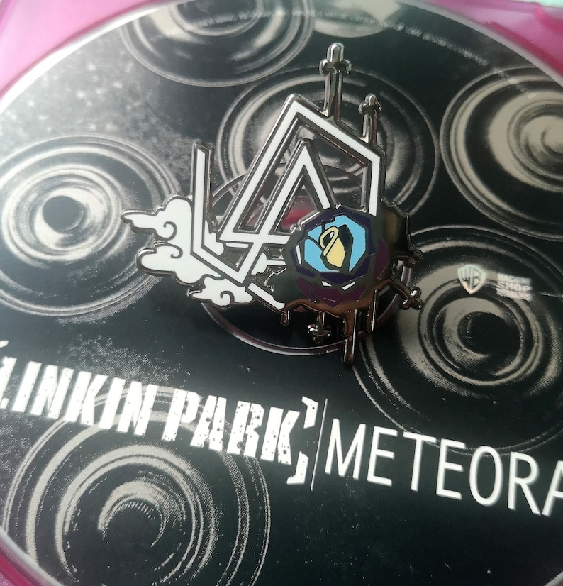 K&ouml;nnte beinhalten: Metall-Anstecknadel mit dem Linkin Park-Logo, einem stilisierten Design einer Wolke und einer geometrischen Form mit einem Krug. Die Anstecknadel befindet sich auf einem schwarzen Hintergrund mit dem Bandnamen und dem Albumtitel "Meteora."