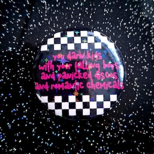 Può includere: Un bottone rotondo con un bordo a scacchi bianchi e neri. Il centro presenta un testo rosa che recita "you dark kids with your falling boys and panicked discos and romantic chemicals". Il bottone ha una finitura lucida.