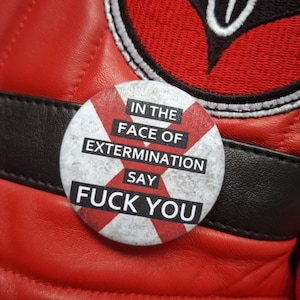 May include: A circular badge with the words "IN THE FACE OF EXTERMINATION SAY FUCK YOU" in black and white. The badge has a red and white striped design. The badge is on a red leather surface.