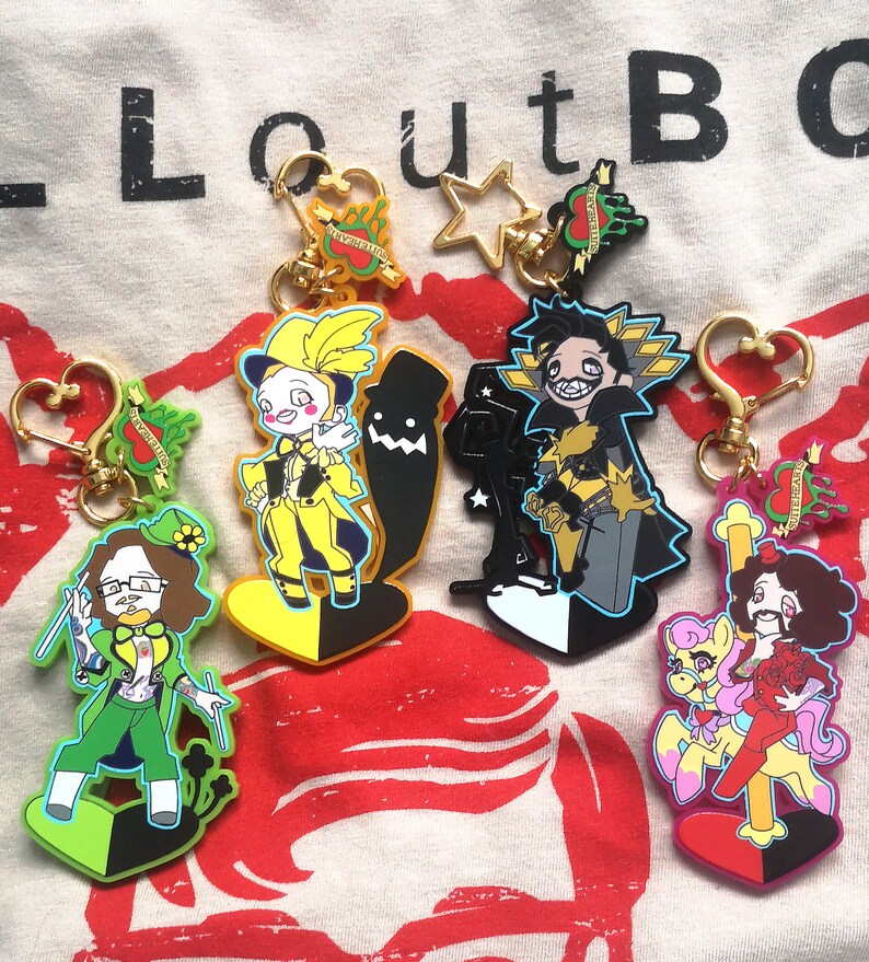 May include: Collection of character keychains with gold-colored hardware. The keychains feature various cartoon figures in vibrant colors, including yellow, green, black, and pink. Some keychains have heart-shaped and star-shaped clasps.