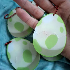 May include: Egg-shaped keyrings with a white and green design. The keyrings have a cracked eggshell appearance with green spots. Each keyring has a red beaded chain and a metal clasp.