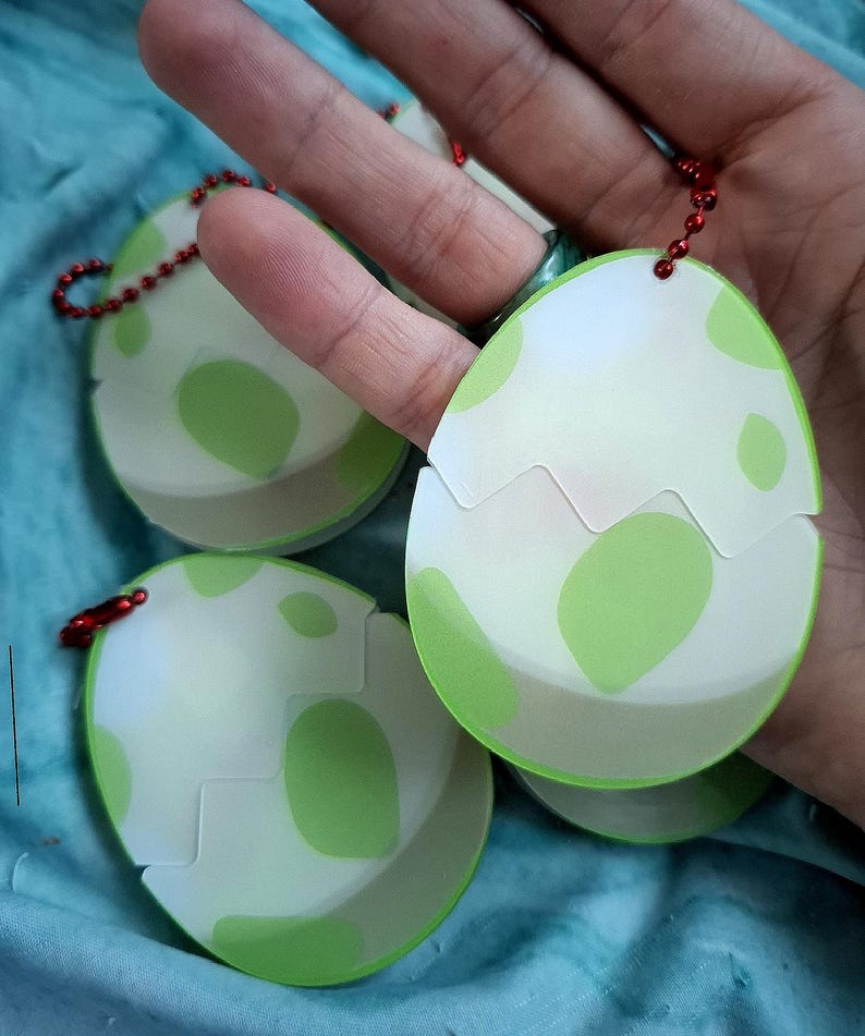 May include: Egg-shaped keyrings with a white and green design. The keyrings have a cracked eggshell appearance with green spots. Each keyring has a red beaded chain and a metal clasp.