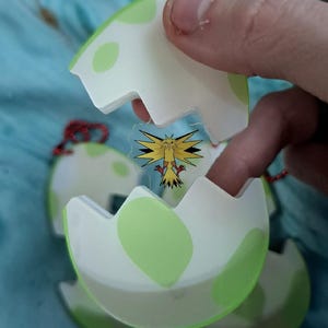May include: A white and green egg-shaped charm featuring a cartoon image of a yellow bird with black and red details. The charm is held by a hand and has a red string attached.