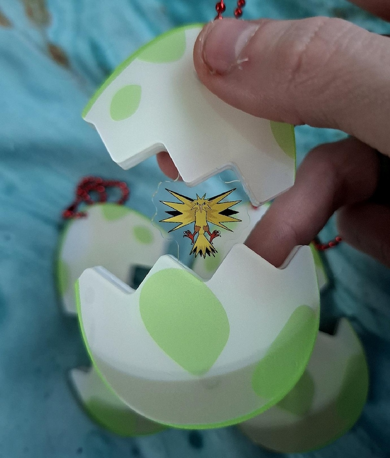 May include: A white and green egg-shaped charm featuring a cartoon image of a yellow bird with black and red details. The charm is held by a hand and has a red string attached.