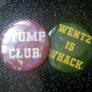 Puede incluir: Dos botones redondos. Uno es rosa con el texto "STUMP CLUB" en blanco. El otro es verde con el texto "WENTZ IS WHACK" en amarillo. Ambos botones tienen un efecto holográfico brillante.