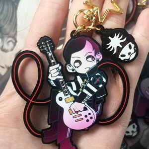 May include: A cartoon keychain depicting a character with black and pink hair playing a pink and white guitar. The character wears a black and white striped jacket. Includes a gold star and skull charm. The keychain is black.