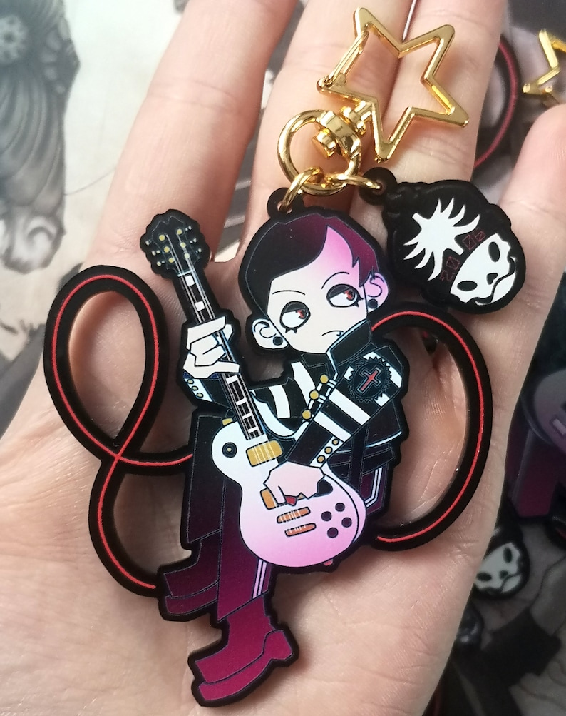 May include: A cartoon keychain depicting a character with black and pink hair playing a pink and white guitar. The character wears a black and white striped jacket. Includes a gold star and skull charm. The keychain is black.