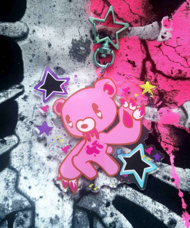 May include: Pink bear keychain with a star-shaped clasp. The bear has a distressed design with blood-like splatters and claw marks. Additional star-shaped charms in black, purple, and yellow are included. The background features a splatter design in pink, black, and white.