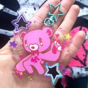May include: A pink bear keychain with a star-shaped clasp. The bear has a cartoonish design with blood-like details. The keychain also features star and heart-shaped accents in various colors, creating a playful and edgy aesthetic.