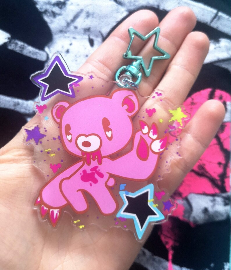 May include: A pink bear keychain with a star-shaped clasp. The bear has a cartoonish design with blood-like details. The keychain also features star and heart-shaped accents in various colors, creating a playful and edgy aesthetic.
