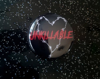 Puede incluir: Un botón redondo negro con la palabra "UNKILLABLE" en rojo. Una forma de corazón está formada por alambre de púas blanco. El botón tiene un acabado brillante.
