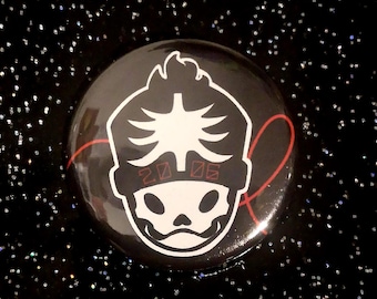 Puede incluir: Un botón negro con un diseño de calavera blanca y el número "2006" en rojo. El botón tiene una línea curva roja en el lado derecho. El diseño es una calavera estilizada con una cresta.
