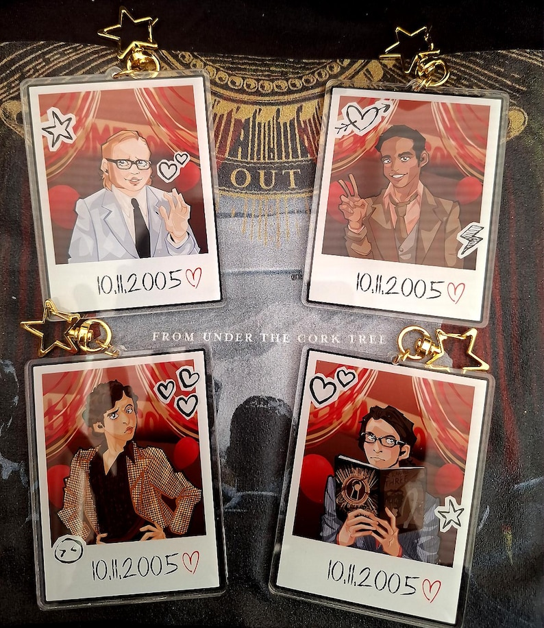 Peut inclure: Quatre porte-cl&eacute;s rectangulaires en acrylique avec des portraits illustr&eacute;s style polaro&iuml;d. Chaque porte-cl&eacute;s poss&egrave;de un fermoir en forme d'&eacute;toile dor&eacute;e et affiche la date 10.11.2005 avec un c&oelig;ur. Les illustrations montrent des personnages et des rideaux rouges.