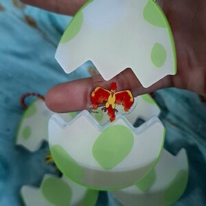 May include: Egg-shaped keyrings with a cartoon bird design. The eggs are white with green spots and a translucent green border. The bird is red and yellow. Each keyring has a red string.