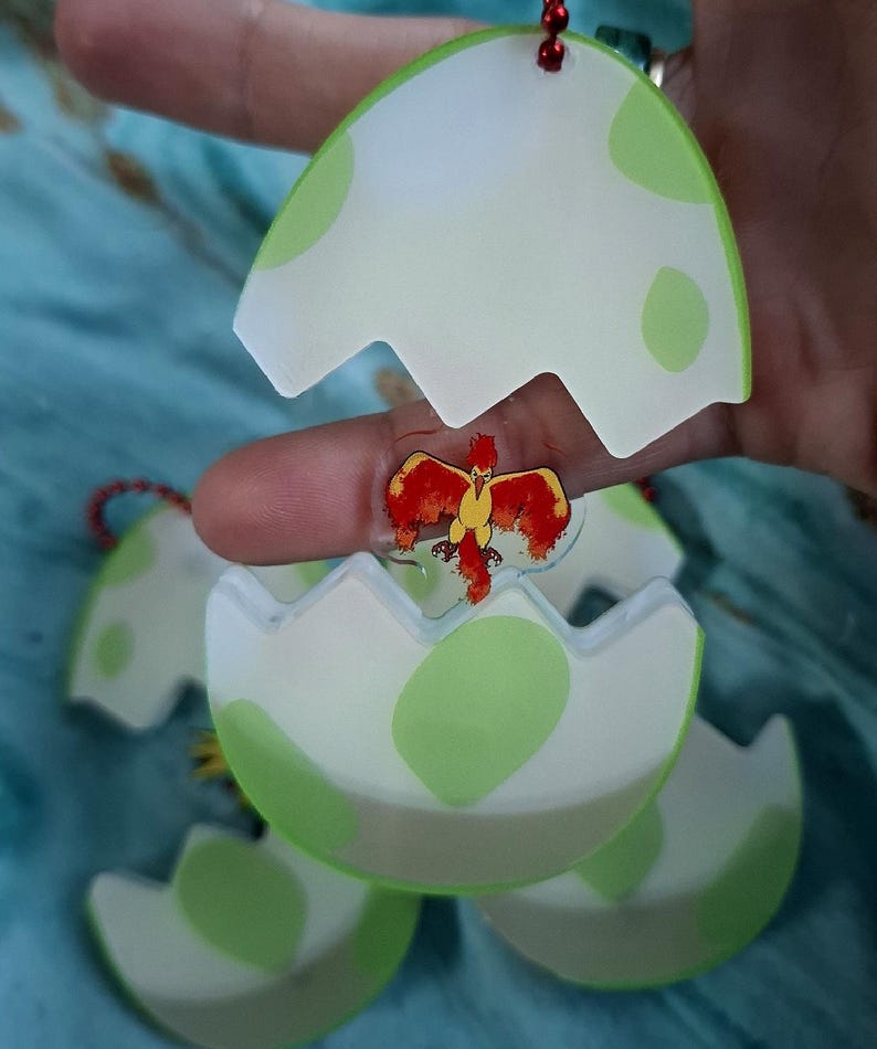 May include: Egg-shaped keyrings with a cartoon bird design. The eggs are white with green spots and a translucent green border. The bird is red and yellow. Each keyring has a red string.