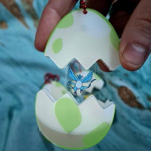 May include: A keyring featuring a green and white egg design, with a cartoon character inside. The egg is open, revealing a blue and white winged creature. The keyring is held by a person.