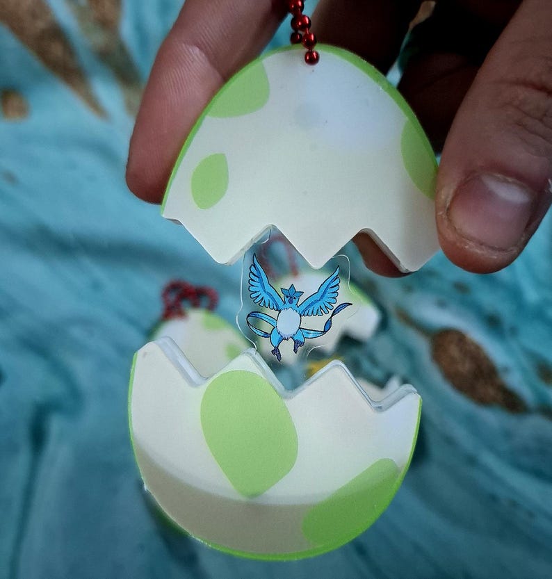 May include: A keyring featuring a green and white egg design, with a cartoon character inside. The egg is open, revealing a blue and white winged creature. The keyring is held by a person.