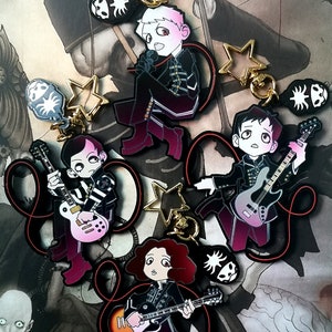 May include: A selection of enamel keyrings featuring cartoon illustrations of band members. Each keyring depicts a musician with a guitar or microphone, in black and burgundy outfits. Additional charms include star shapes and a skull design.