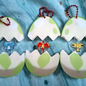 May include: Six egg-shaped keyrings with a white and green design, each with a small charm of a cartoon creature. The keyrings have a red beaded chain for attachment. The eggshells are open, revealing the creatures.