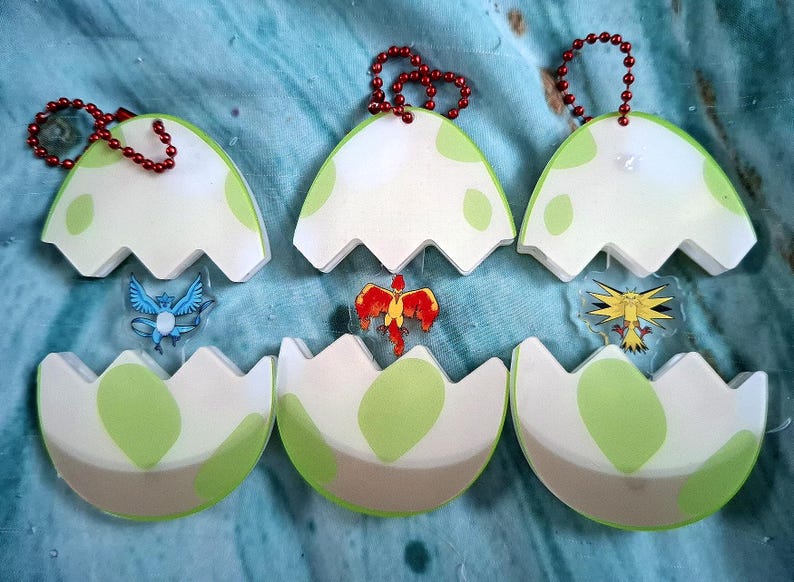 May include: Six egg-shaped keyrings with a white and green design, each with a small charm of a cartoon creature. The keyrings have a red beaded chain for attachment. The eggshells are open, revealing the creatures.