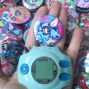 May include: A light blue Digivice toy with a digital screen and buttons, held with a round button featuring a blue and pink cartoon dragon. Many other colourful buttons with similar designs are in the background.