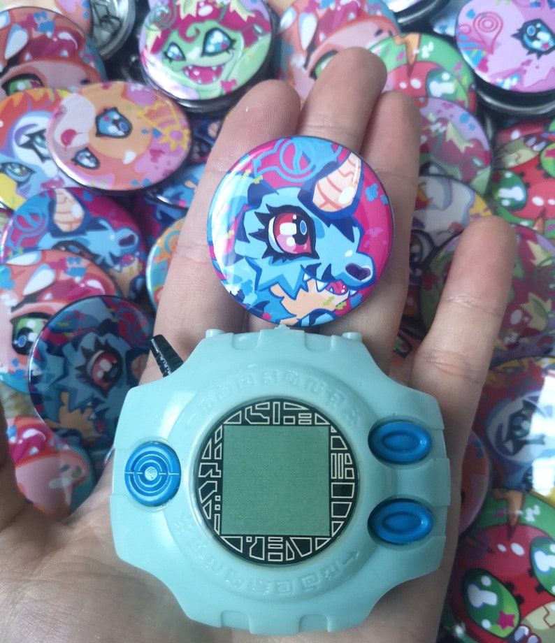 May include: A light blue Digivice toy with a digital screen and buttons, held with a round button featuring a blue and pink cartoon dragon. Many other colourful buttons with similar designs are in the background.