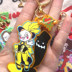 May include: A yellow and black keychain featuring a cartoon character in a yellow outfit and a black silhouette. The keychain has a gold-colored heart-shaped clasp and a small tag that says "SUITEHEARTS".