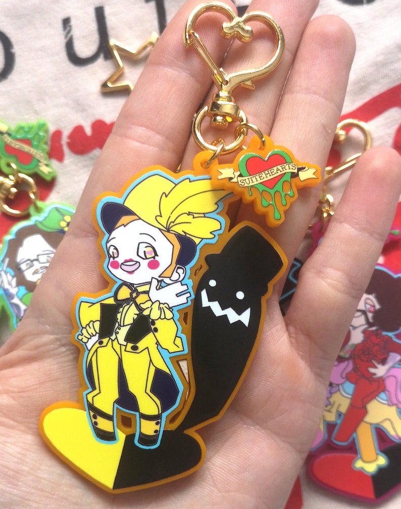 May include: A yellow and black keychain featuring a cartoon character in a yellow outfit and a black silhouette. The keychain has a gold-colored heart-shaped clasp and a small tag that says "SUITEHEARTS".