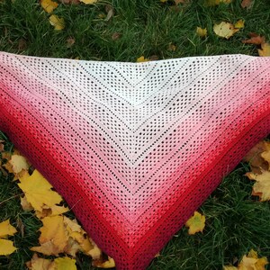 May include: A triangular crocheted shawl with a gradient of red, pink, and white. The shawl is laid out on a bed of green grass with fallen autumn leaves.