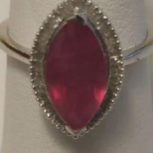 May include: A silver ring featuring a marquise-cut, deep pink gemstone. The gemstone is surrounded by a beaded border. The ring is displayed on a white surface.