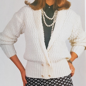 Puede incluir: Cárdigan de punto color crema con un patrón texturizado. El suéter de doble botonadura tiene botones grandes y un dobladillo acanalado. Las mangas son largas y el cuello es estilo chal. El cárdigan se combina con una falda estampada en blanco y negro.