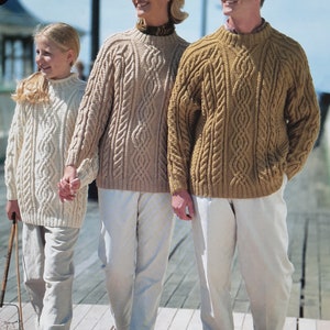 May include: Three people wearing cable knit sweaters in different colors: beige, white, and brown. The sweaters have a traditional cable knit pattern. The people are standing on a wooden boardwalk.