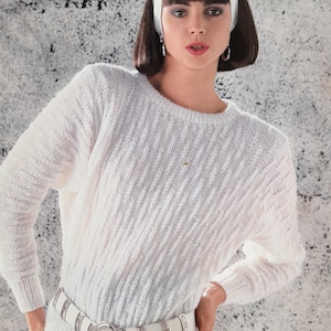May include: White knitted sweater with a white belt and a silver buckle. The sweater has a cable knit pattern. The model is wearing a white headband with a pearl accent.