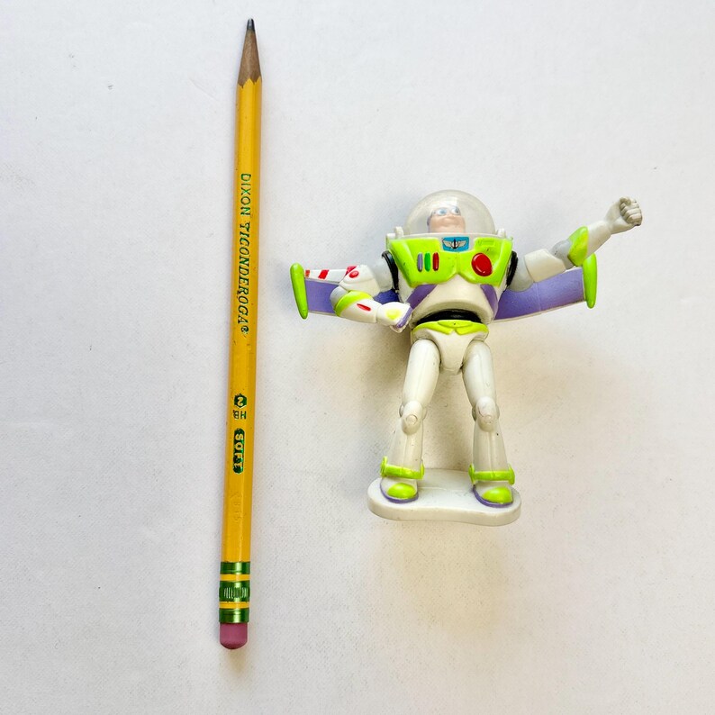 May include: A plastic Buzz Lightyear action figure with a clear helmet, white, green, and purple suit, and extended wings, positioned next to a yellow Dixon Ticonderoga number 2 pencil for scale, showing the figure is approximately 4 inches tall.