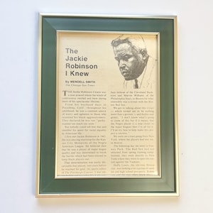 May include: A framed newspaper clipping titled The Jackie Robinson I Knew by Wendell Smith from The Chicago Sun Times. The print features a black and white sketch of Jackie Robinson and columns of text detailing his life and career history.