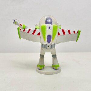 May include: A plastic action figure of a space ranger character featuring a white, green, and purple color scheme. The figure has extended wings with red and white diagonal stripes, a clear dome helmet, and stands on a circular base.