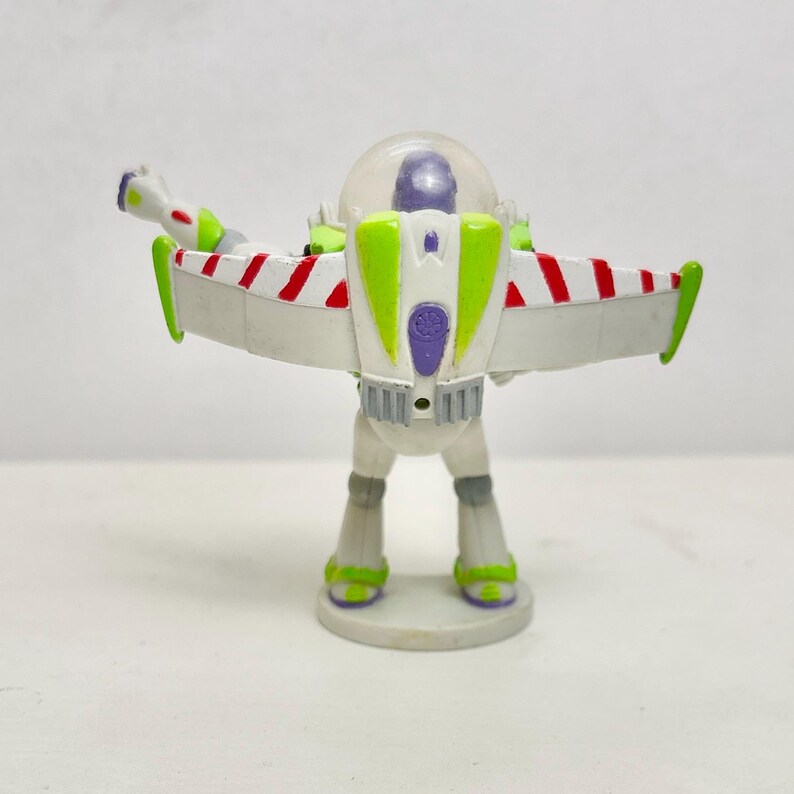 May include: A plastic action figure of a space ranger character featuring a white, green, and purple color scheme. The figure has extended wings with red and white diagonal stripes, a clear dome helmet, and stands on a circular base.