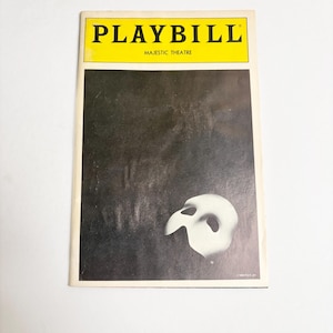 May include: A Playbill program for the Majestic Theatre featuring a black background with a white theatrical mask in the lower right corner. The top section has a yellow banner with the text PLAYBILL in black and MAJESTIC THEATRE in smaller text.