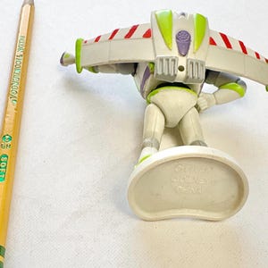 May include: A Buzz Lightyear action figure with white, green, and purple plastic parts, featuring wings with red and white stripes. The figure stands on a white base marked with Pixar, Disney, and China. A yellow Dixon Ticonderoga pencil is visible.