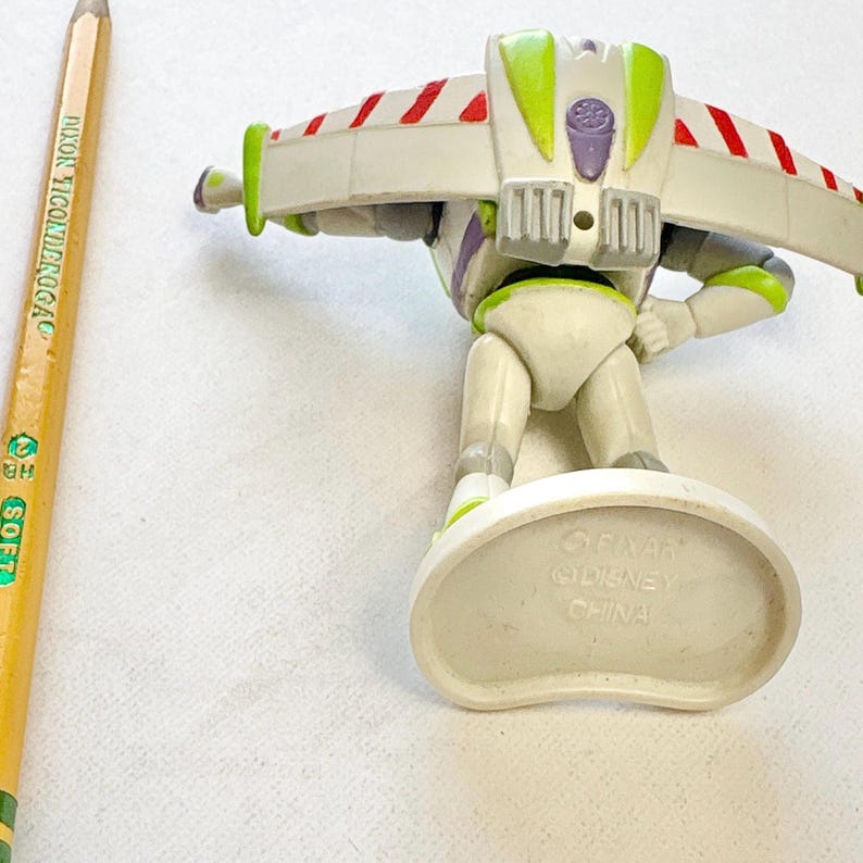 May include: A Buzz Lightyear action figure with white, green, and purple plastic parts, featuring wings with red and white stripes. The figure stands on a white base marked with Pixar, Disney, and China. A yellow Dixon Ticonderoga pencil is visible.