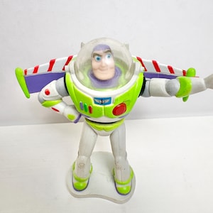 May include: A Buzz Lightyear action figure featuring a white, lime green, and purple space suit with a clear plastic helmet dome. The figure has extended wings with red and white stripes, a red circular button, and a blue chest emblem.