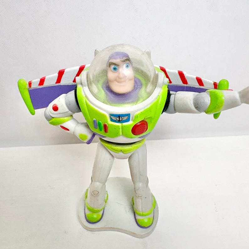 May include: A Buzz Lightyear action figure featuring a white, lime green, and purple space suit with a clear plastic helmet dome. The figure has extended wings with red and white stripes, a red circular button, and a blue chest emblem.