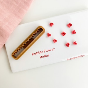 Può includere: Uno strumento di rotolamento per fiori a bolle in plastica marrone su una superficie bianca con nove fiori a bolle rossi sparsi intorno. Il testo "Bubble Flower Roller" e "@creativewellco" è stampato sulla superficie.
