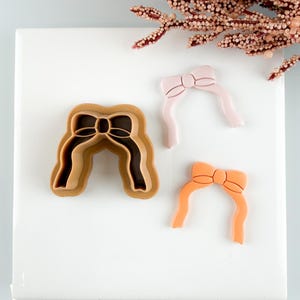 Könnte beinhalten: Drei Ausstecher in Schleifenform und zwei Schleifen-Dekorationen werden auf einer weißen Oberfläche gezeigt. Die Ausstecher sind in Brauntönen gehalten, während die Dekorationen hellrosa und orange sind. Ein dekorativer Blumenstrauß befindet sich im Hintergrund.