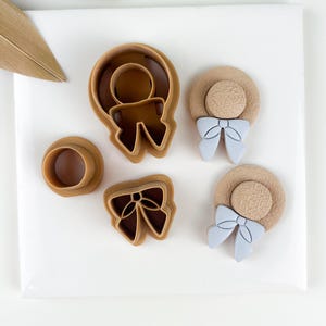 Può includere: Una collezione di formine per biscotti in plastica marrone di varie forme, tra cui una persona, un fiocco e un cappello con un fiocco. Le formine a cappello hanno fiocchi azzurri. Disposti su una superficie bianca.