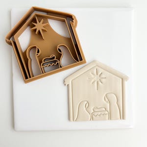 May include: A brown plastic cookie cutter in the shape of a nativity scene with a star above the stable. The cutter is shown next to a white clay cutout of the same design.
