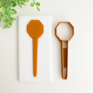 May include: Two brown plastic octagon-shaped cookie cutters on a white background. The cutter on the left is solid, while the cutter on the right is hollow.