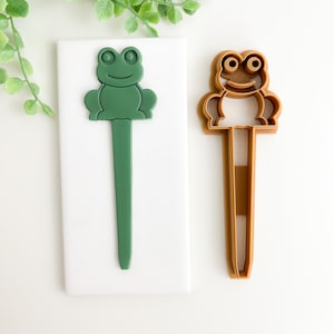 May include: Two frog-shaped cookie cutters. One is a solid green color, and the other is a brown outline. Both have a long, thin handle. The frog's face has large eyes and a smiling mouth. The cutters are on a white surface.